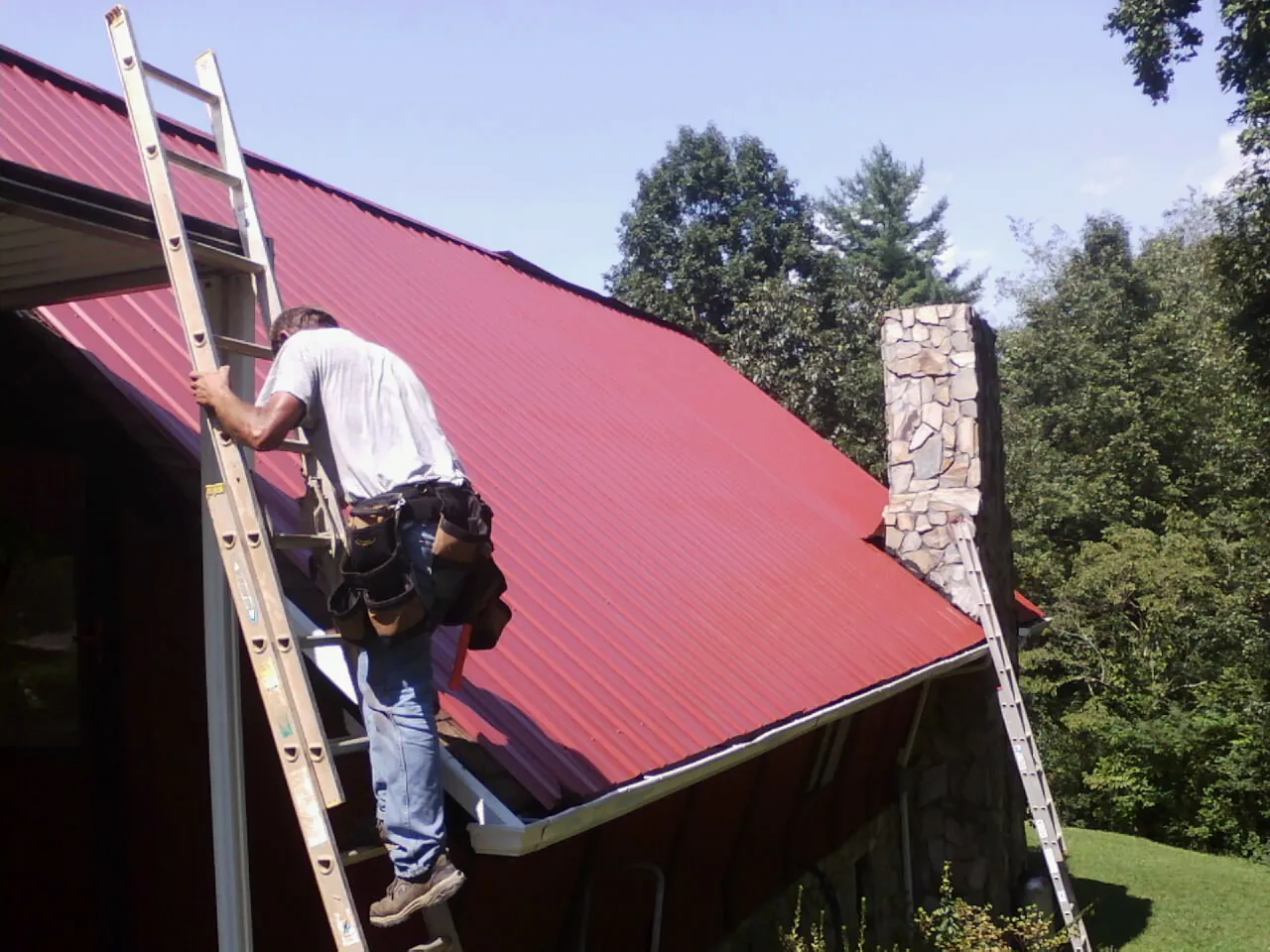 Roofer on a ladder beside a red metal roof and stone chimney — Soffit & Fascia Repair by Roofing Brothers in Peach Bottom